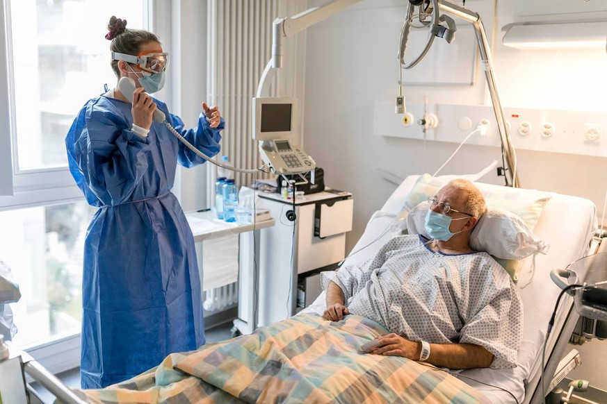 Pflegefachfrau Nora Tschanz benutzt das Telefon, um sich etwas bringen zu lassen, statt das Zimmer verlassen und sich umziehen zu müssen. Covid-Patient Roland Lerch trägt solange eine Hygienemaske.