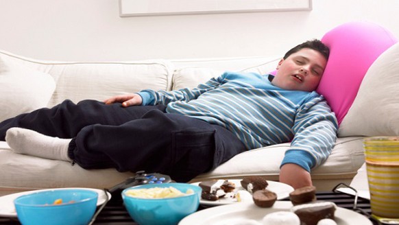Young, Overweight Boy Sleeps on a Sofa Next to a Table of Crisps and Biscuits