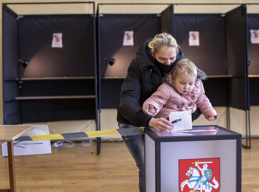 A woman wearing a face mask to protect against the coronavirus, with a child, casts her ballot at a polling station during the second round of a parliamentary election in Vilnius, Lithuania, Sunday, O ...