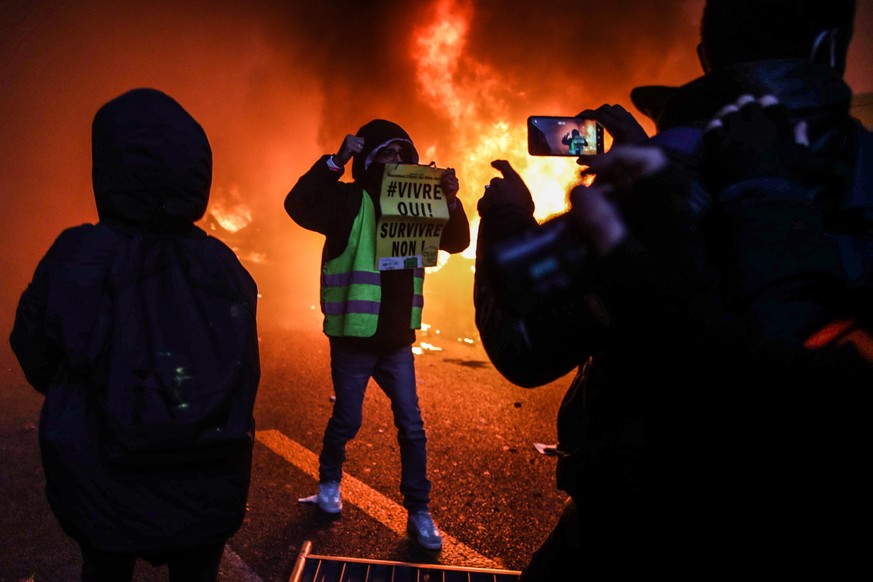 epa08864960 An activist from Gilet Jaune (Yellow Vests) holds a sign says &#039;Live yes, survive no&#039; as several cars are on fire during a protest against France&#039;s controversial global secur ...