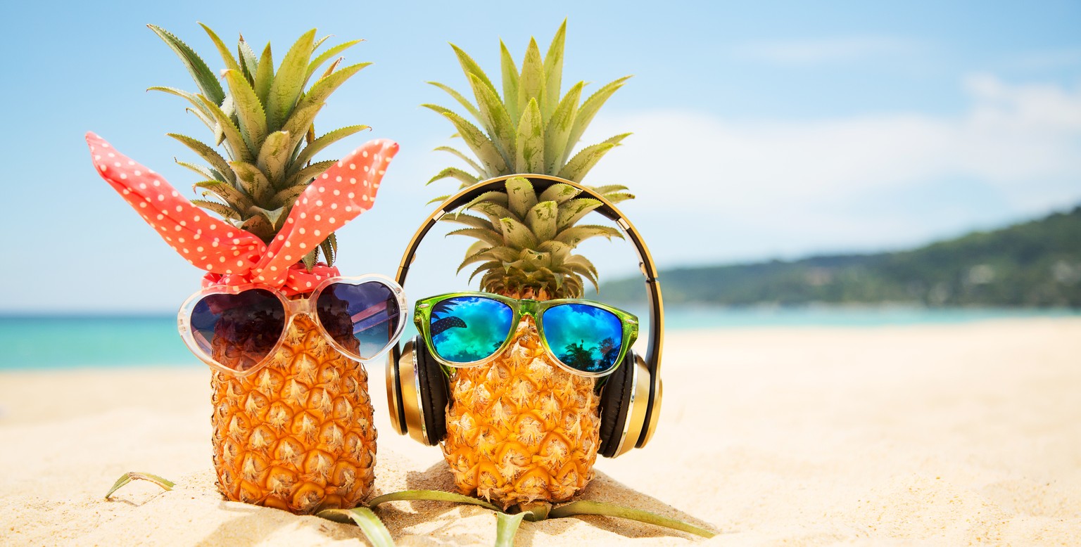 Couple of attractive pineapples in love on the sand against turquoise sea. Wearing stylish mirrored sunglasses. Tropical summer vacation concept. Sunny day on the beach of tropical island. Honeymoon