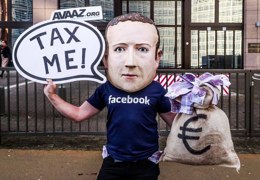 epa07207687 An activist wearing a mask depicting Facebook&#039;s CEO Mark Zuckerberg holds a banner reading &#039;Tax me&#039; at the start of an European Union Finance Ministers Meeting in front of t ...