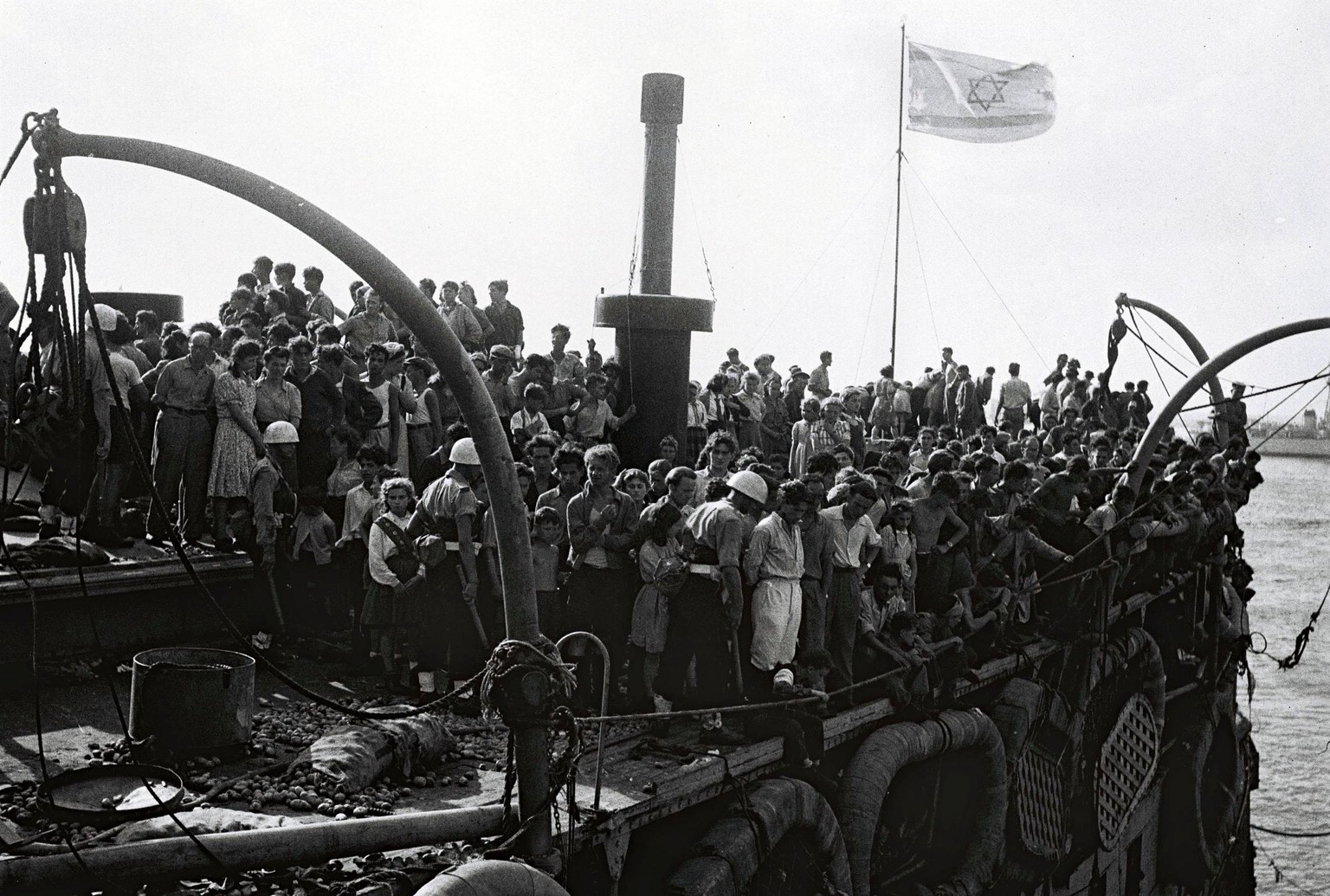 HAIFA, MANDATE PALESTINE - JULY 18, 1947: The Israeli flag flies over the crowded upper deck of the illegal immigration ship Exodus, carrying Jewish refugees from war-torn Europe, July 18, 1947 before ...