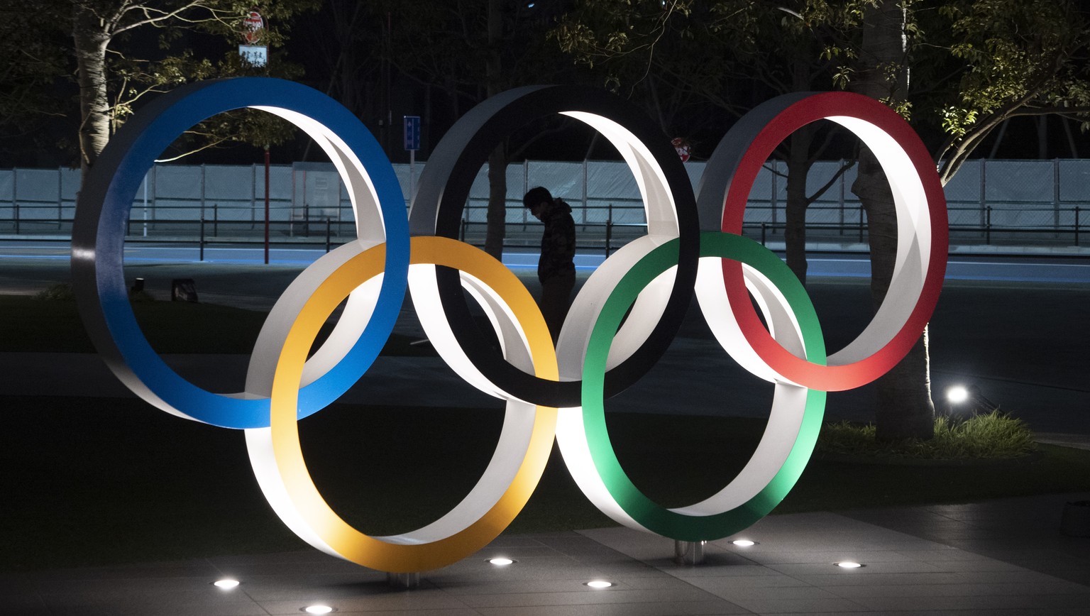 FILE - In this March 24, 2020, a man is seen through the Olympic rings in front of the New National Stadium in Tokyo. IOC officials say the Tokyo Olympics will open on July 23 and almost nothing now c ...