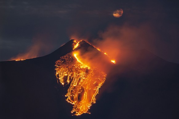The moon is partially seen in the sky as lava flows from the Mt Etna volcano, near Catania in Sicily, southern Italy, early Tuesday, Feb. 23 , 2021. The explosion started before midnight on Monday nig ...