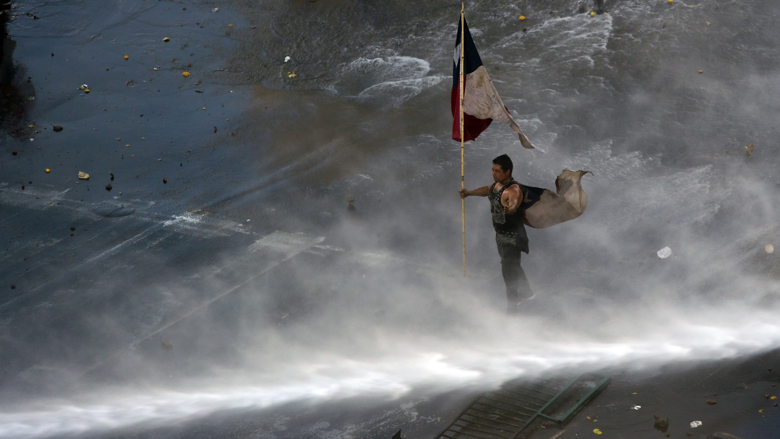 Holding a Chilean flag, a protester braces against a water canon sprayed from a police truck amid a march by students and union members in Santiago, Chile, Monday, Oct. 21, 2019. Protesters defied an  ...