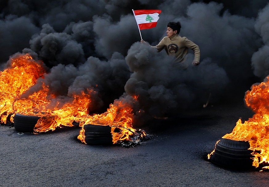 An anti-government demonstrator jumps on tires that were set on fire to block a main highway as he holds a national flag, during a protest in the town of Jal el-Dib, north of Beirut, Lebanon, Tuesday, ...