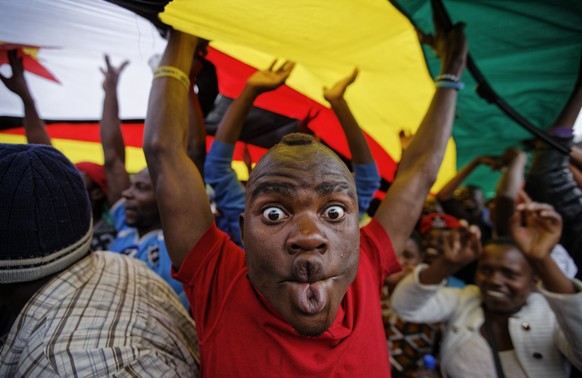 A happy protester pulls a face as he and others stand under a large national flag, at a demonstration of tens of thousands at Zimbabwe Grounds in Harare, Zimbabwe Saturday, Nov. 18, 2017. Opponents of ...