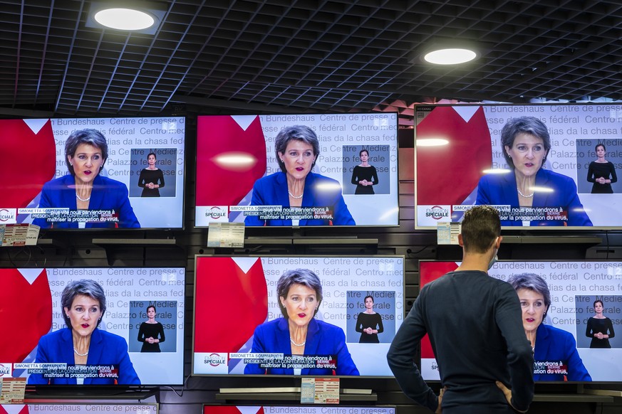 A man wearing a protective mask watches Swiss President Simonetta Sommaruga as she gestures during a press conference in Bern on TV screens inside a showroom at the Fnac store announcing new measures  ...