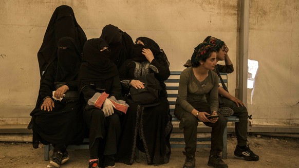 Women related to fighters of the Islamic State group sit next to Syrian Democratic Forces guards as they wait to board buses and trucks, leaving the overcrowded a an overcrowded al-Hol camp to return  ...