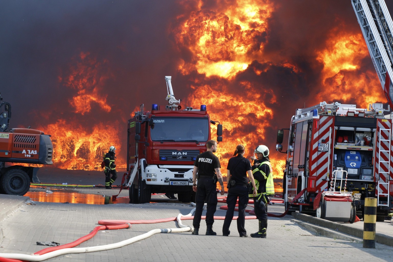 30.05.2018, Mecklenburg-Vorpommern, Rostock: Einsatzkraefte der Polizei und Feuerwehr stehen bei einem Brand auf dem Gelaende eines Entsorgungsunternehmens im Stadtteil Neu Hinrichsdorf vor einer Feue ...
