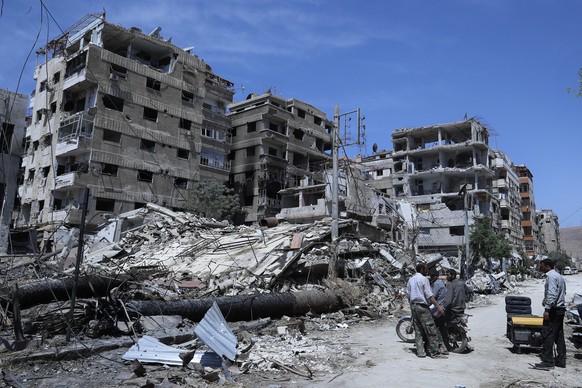FILE - In this April 16, 2018, file photo, people stand in front of damaged buildings, in the town of Douma, near Damascus, Syria. In its ninth year, many Syrians fear their countryÄôs unresolved war ...
