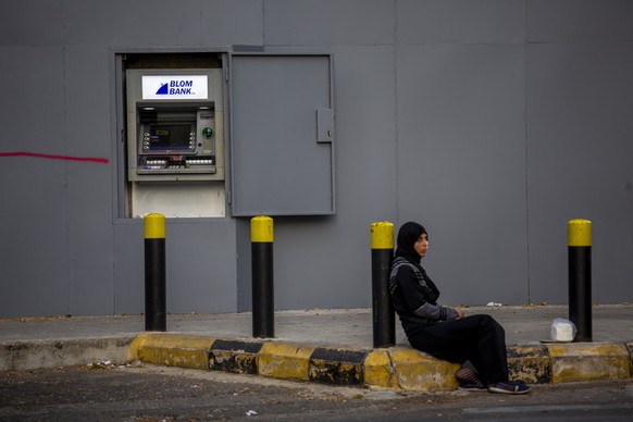 In this Thursday, May 21, 2020 photo, a woman begs on a sidewalk in front an ATM machine covered by iron shields along the facade of a bank to prevent acts of sabotage, in Beirut, Lebanon.Riad Salameh ...